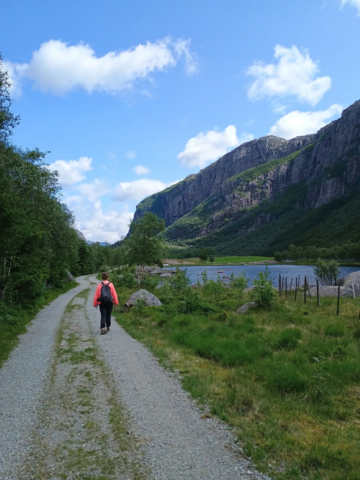 Wandelaar in roze jas op een grindpad langs een bergmeer met steile bergwanden op de achtergrond in Rogaland.