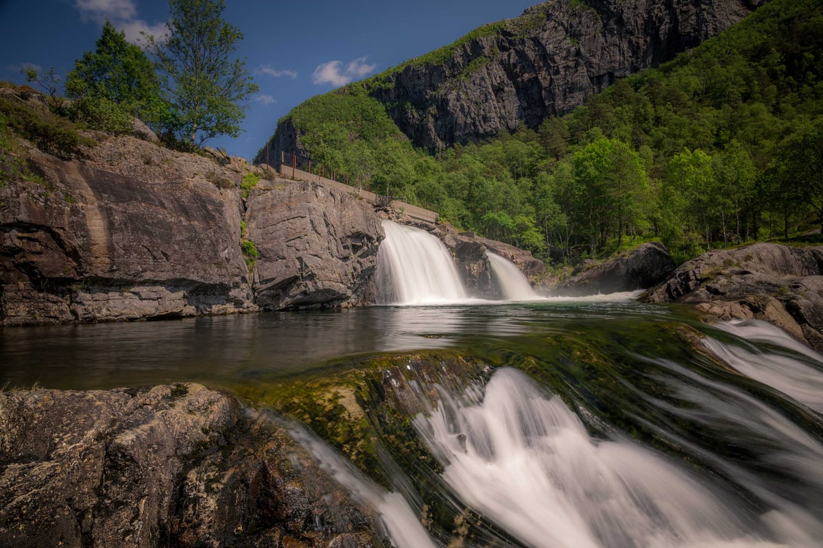 Tweedelige waterval stroomt over grijze rotsen in een groene bergvallei op een zonnige zomerdag.