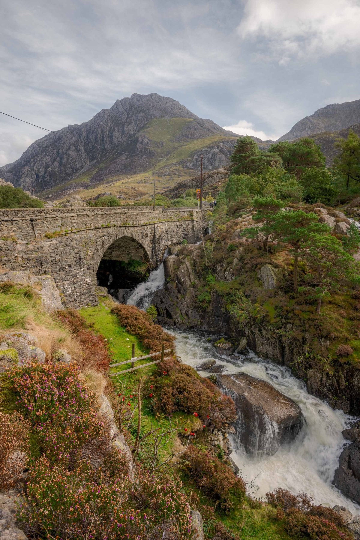 Stenen boogbrug over waterval met bergen op de achtergrond.