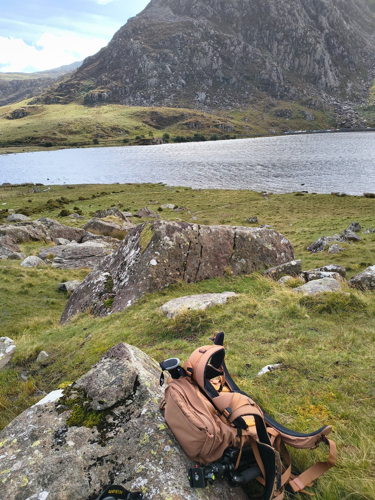 Bruine camera-rugzak met Nikon-camera op een rotsblok met korstmos, met Llyn Ogwen en rotswanden op de achtergrond