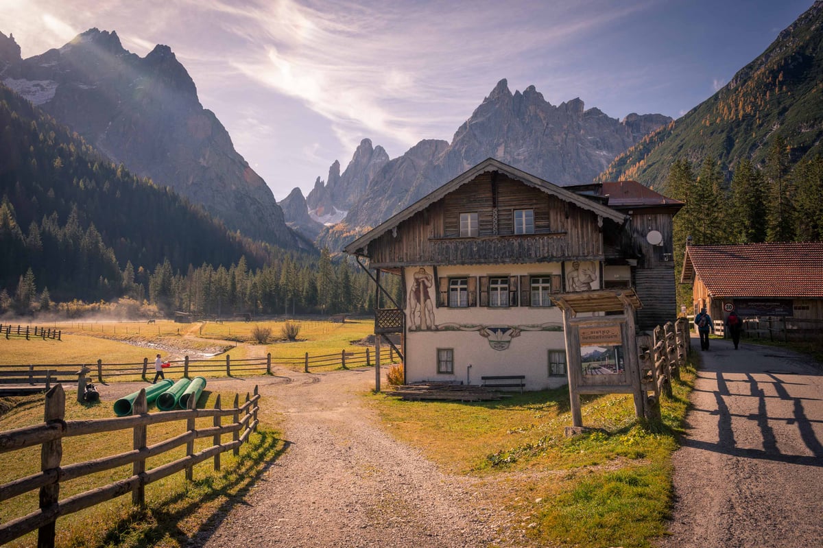 Traditioneel beschilderd Alpenhuis aan een onverharde weg in een breed bergdal met dramatische Dolomieten-pieken op de achtergrond.