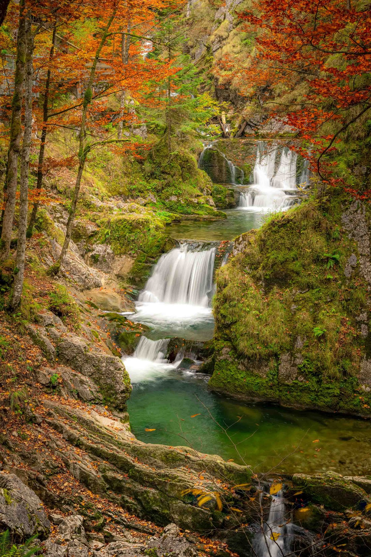 Meervoudige waterval met zijdezacht stromend water in een herfstbos met rode bladeren en smaragdgroene poelen.