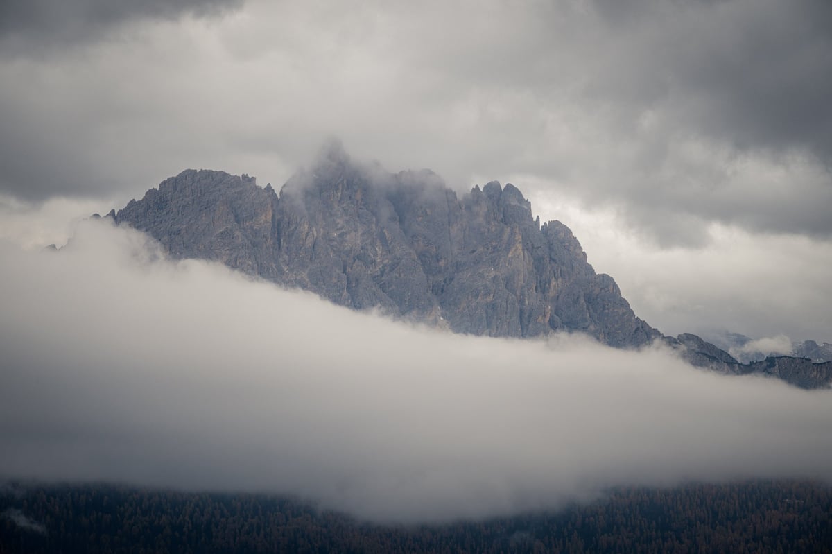Spits rotsachtig bergmassief omgeven door lage wolken en een bewolkte grijze lucht, met naaldbos zichtbaar aan de voet.