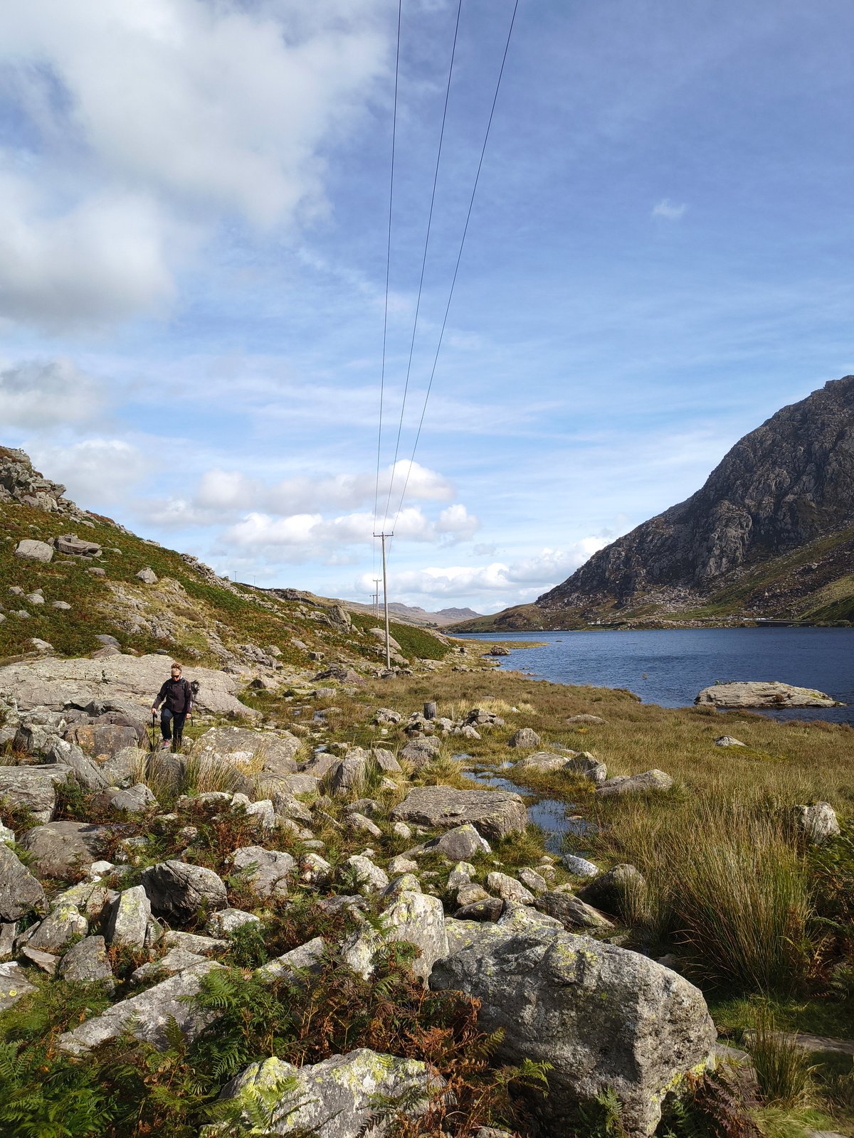 Wandelaar met trekstokken op een rotsachtig pad langs het donkerblauwe water van Llyn Ogwen, steile rotswanden rechts