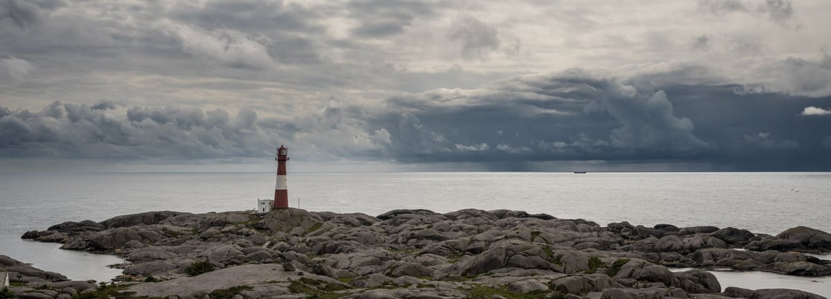 Rood-witte vuurtoren op een rotsachtig eiland onder een dreigende bewolkte hemel boven een kalme zee.