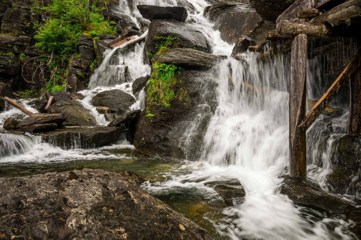Meervoudige waterval die over donkere rotsblokken stroomt met oude houten balken op de voorgrond bij het molenhuis van Jonegarden