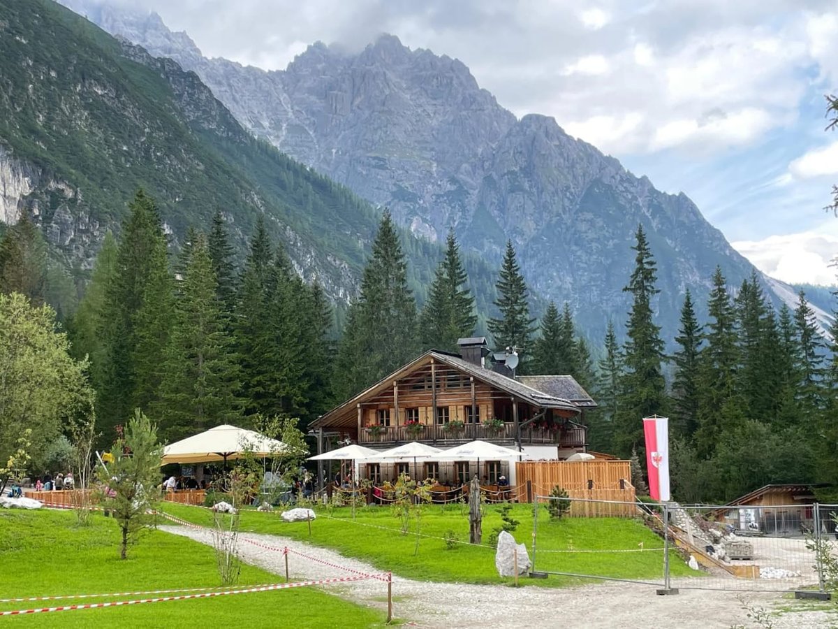 Traditionele Alpenhut met terras in een groen bergdal, met ruige rotstoppen op de achtergrond.