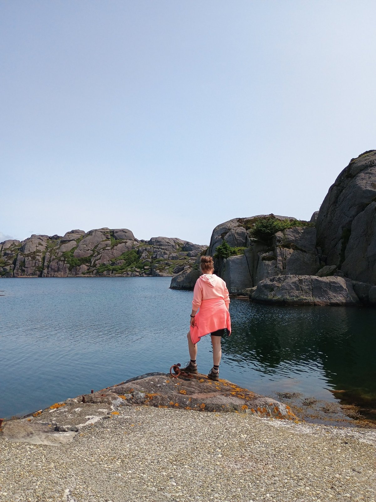 Vrouw in roze hoodie staat op een rots en kijkt uit over een rustige baai met granieten rotsen op de achtergrond.
