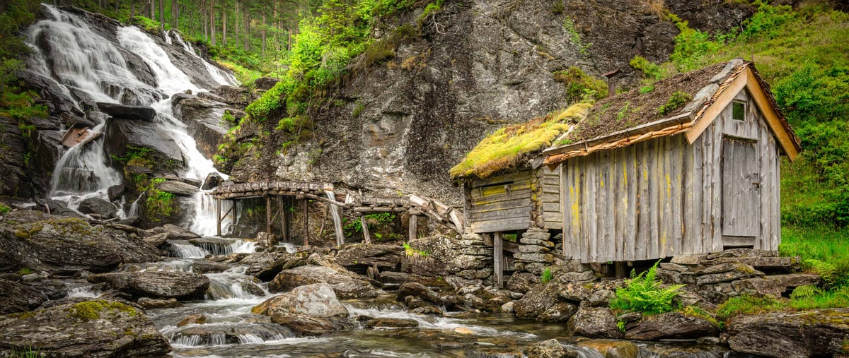 Verlaten houten molen op stenen pijlers naast een getrapte waterval in een groen Scandinavisch berglandschap bij Jonegarden