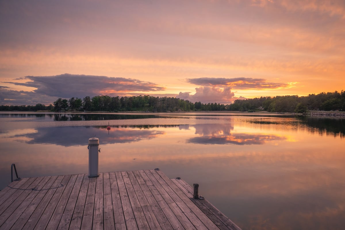 Houten steiger uitkijkend over een spiegelglad meer bij een kleurrijke oranje-roze zonsondergang met bosrijke oever op de achtergrond.