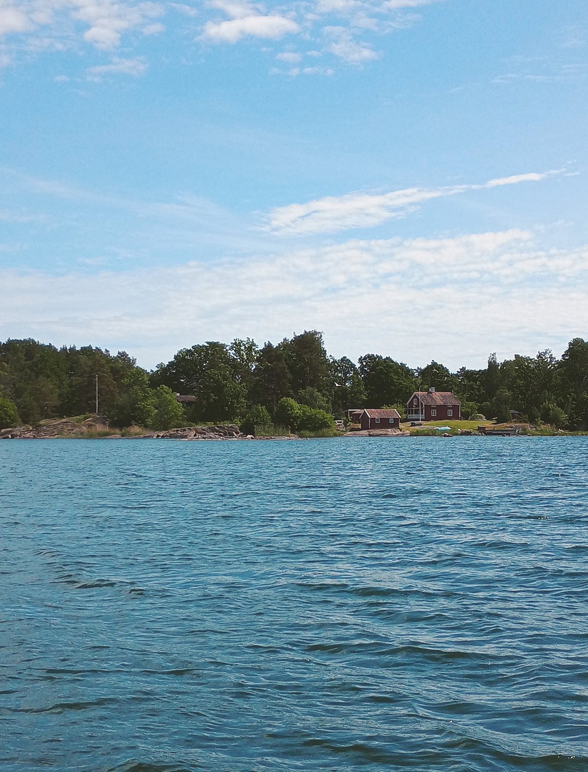 Rood Scandinavisch houten huisje aan een rotsige bosrijke oever, gefotografeerd vanaf het water op een zonnige zomerdag.