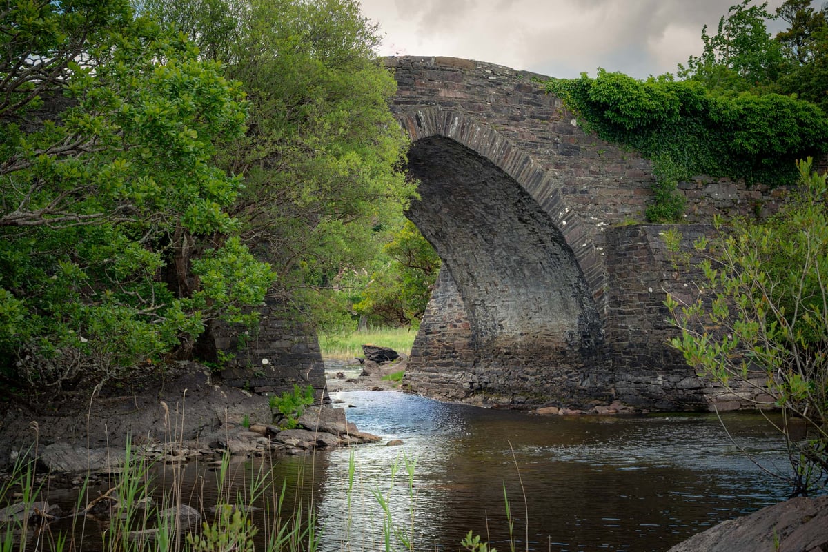 Oude stenen boogbrug over een rotsige rivier omgeven door groen gebladerte bij Muckross Lake