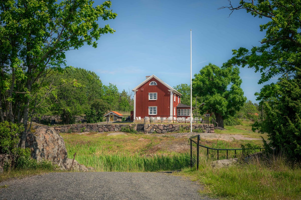 Traditioneel rood Zweeds houten huis met witte kozijnen op een rotsachtig terrein, omgeven door groene bomen in de zomer.