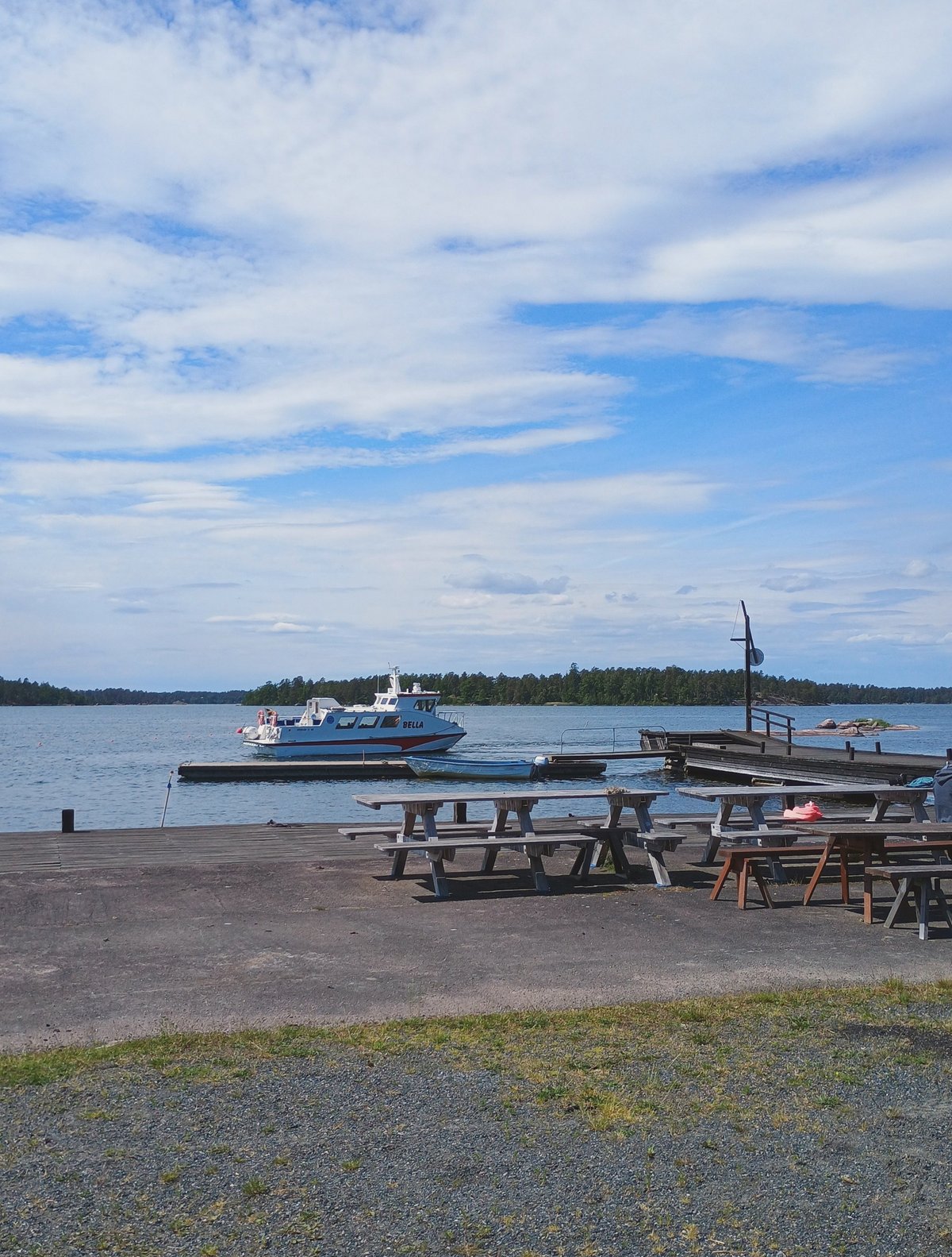 Houten picknicktafels aan een steiger met een aangemeerde motorboot genaamd Bella en een bosrijke archipelago op de achtergrond.