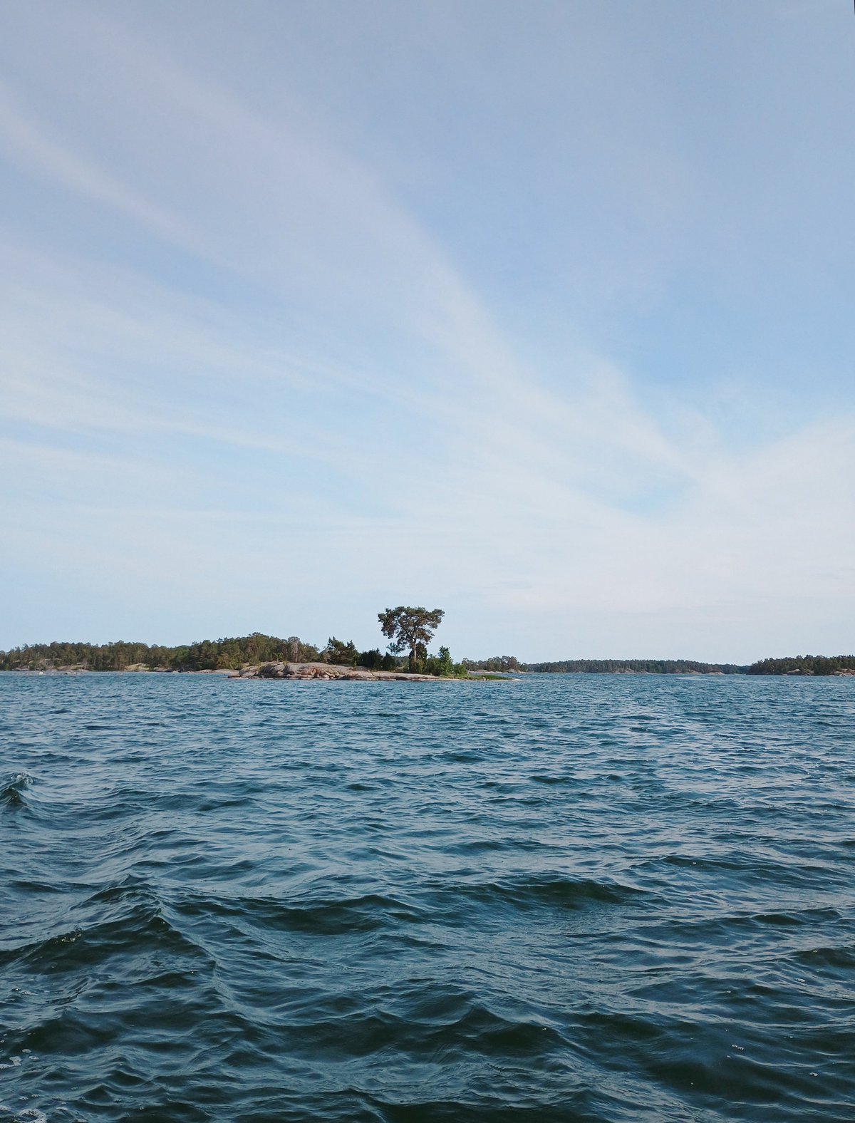 Klein rotsachtig eilandje met één solitaire boom omgeven door blauw water in een archipel, onder een lichtblauwe zomerlucht.