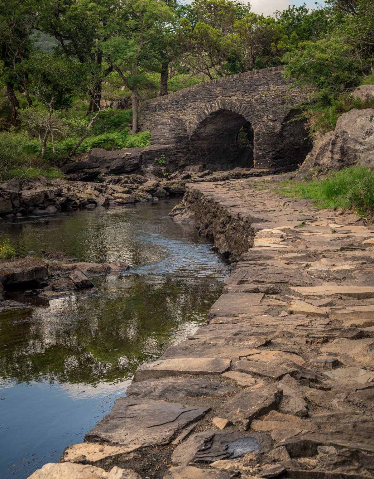 Historische stenen boogbrug over een beek bereikbaar via een oud leisteen klinkerpad omringd door groen