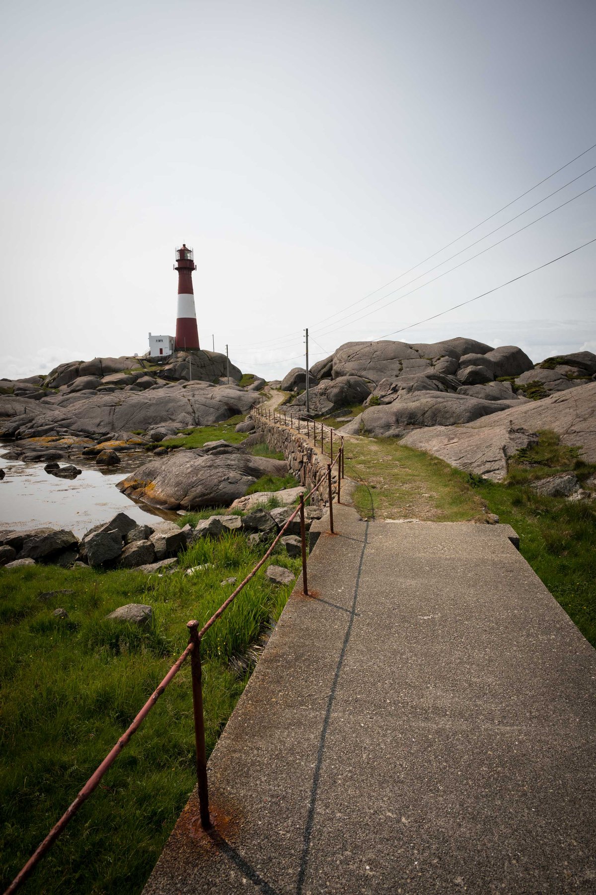 Betonnen pad met roestige leuningen leidt naar een rood-witte vuurtoren op een rotsachtig Noors eiland.