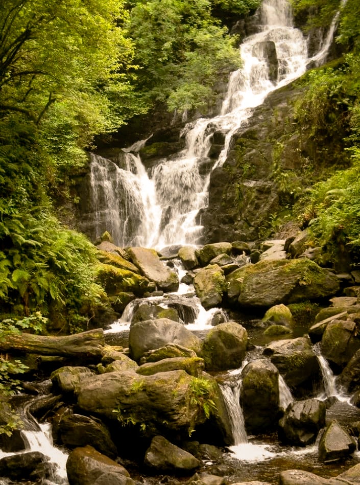 Meervoudige waterval die in trappen over bemost gesteente stroomt in groen loofbos bij Torc Waterfall