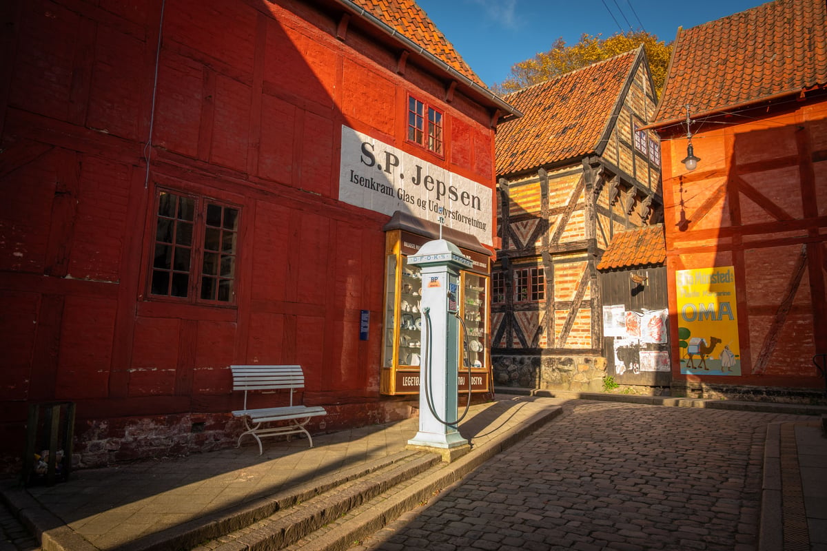 Historisch Deens plein met rode vakwerkgebouwen, een vintage benzinepomp en een winkelgevel van S.P. Jepsen in een openluchtmuseum.