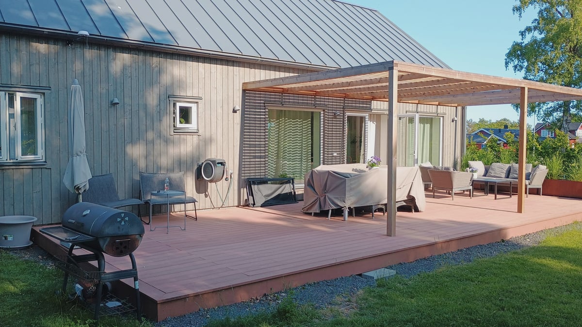 Houten vlonderterras met pergola en loungemeubilair bij een Scandinavisch huis op een zonnige zomerdag.