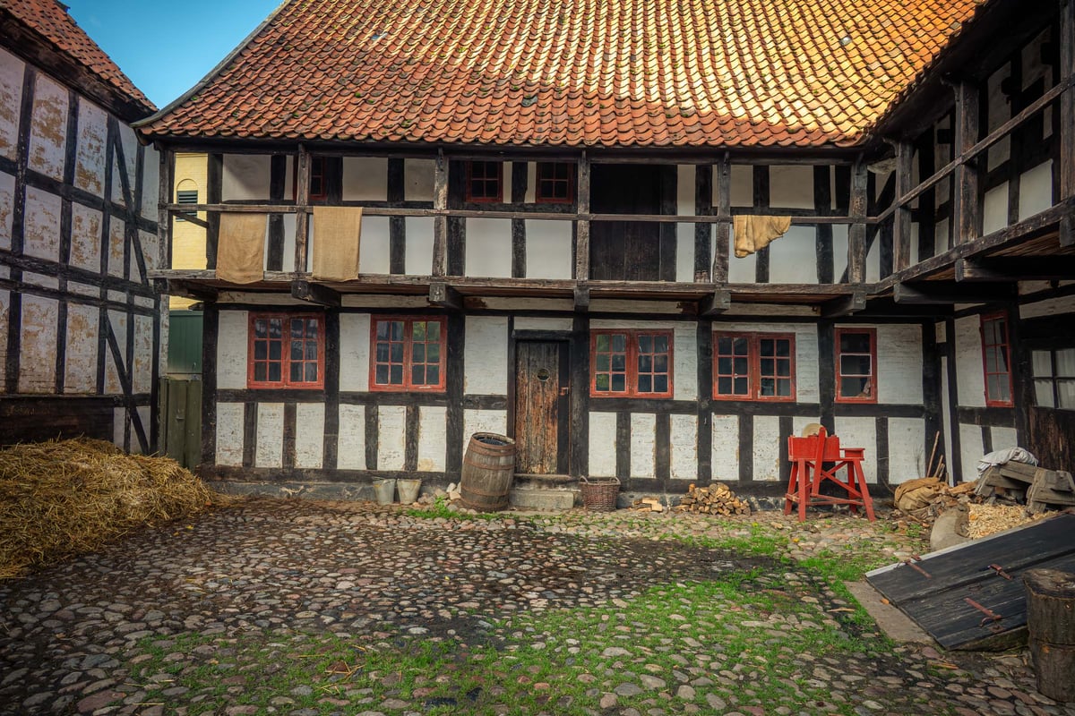 Binnenplaats van een historisch vakwerkgebouw met keienbestrating, hooibalen, een houten vat en rode raamkozijnen.