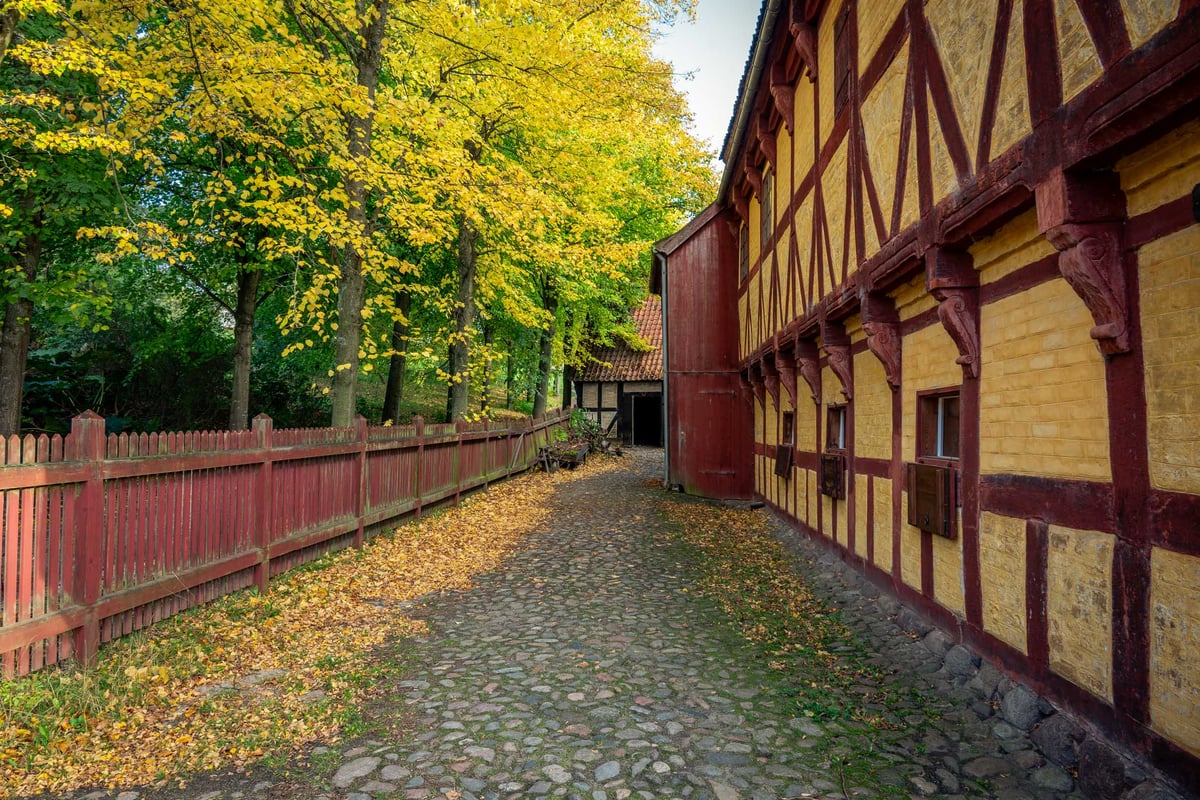 Kasseienpad tussen een historisch rood-geel vakwerkgebouw en een rode houten omheining, omgeven door herfstbomen.