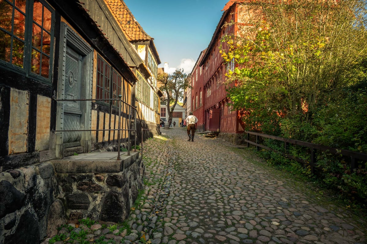 Een historisch geplaveid steegje met vakwerkhuizen en een rode schuur, met een figuur in historische klederdracht die wegloopt.