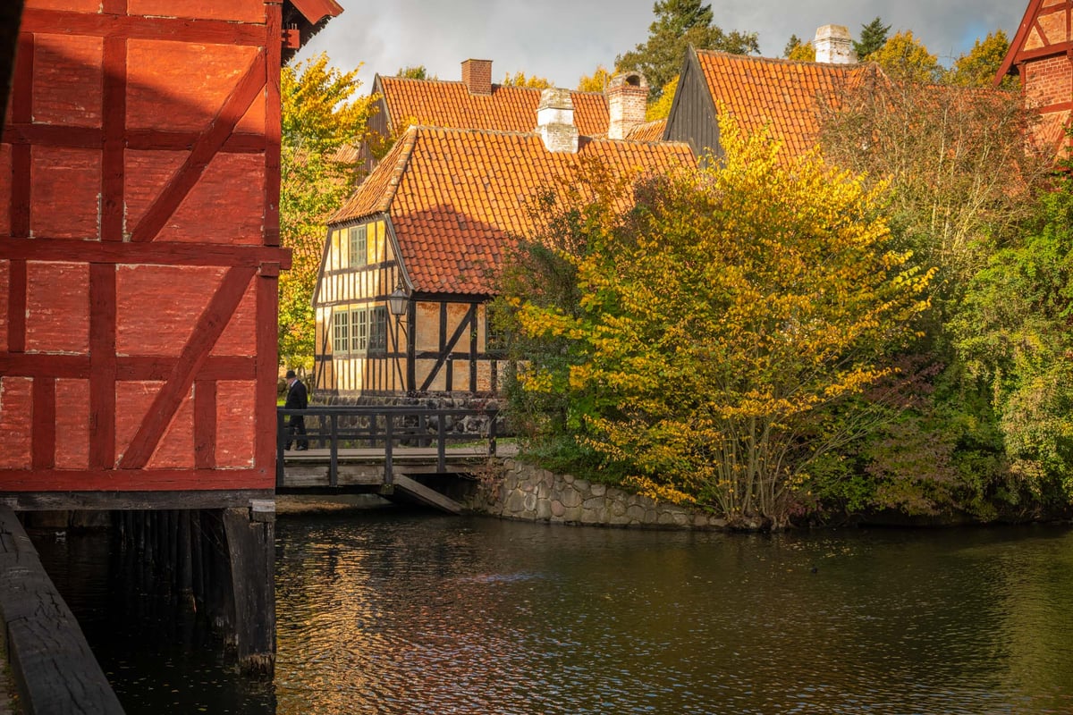Roodgeschilderd vakwerkhuis op palen langs een waterpartij met herfstbomen en traditionele Scandinavische gebouwen op de achtergrond.