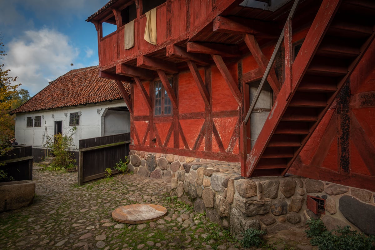 Rood vakwerkhuis met houten buitentrap en stenen fundering op een kinderkopjesplein, vermoedelijk een Scandinavisch openluchtmuseum.
