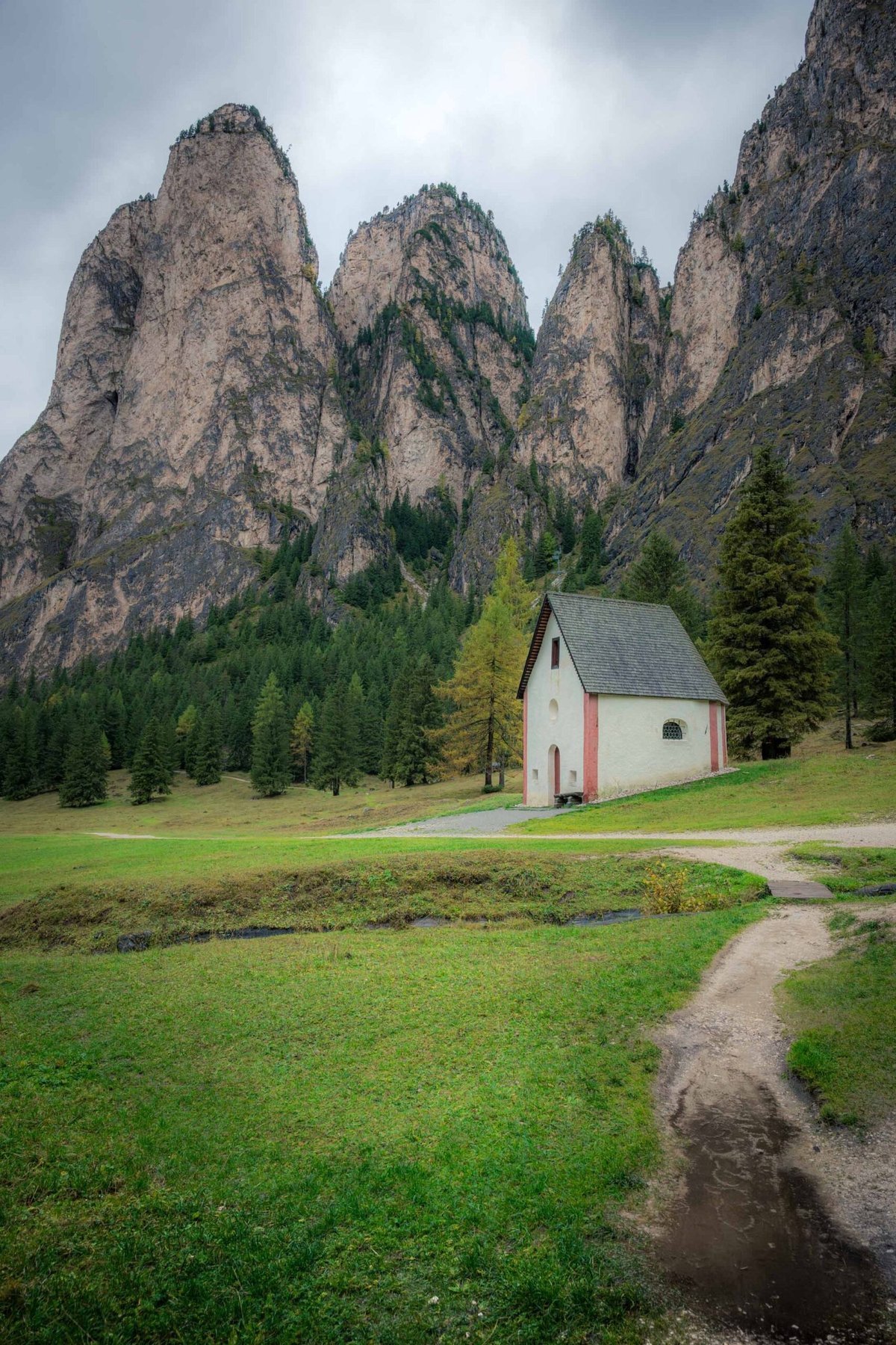 Small white chapel in green Alpine valley beneath dramatic Dolomite peaks.