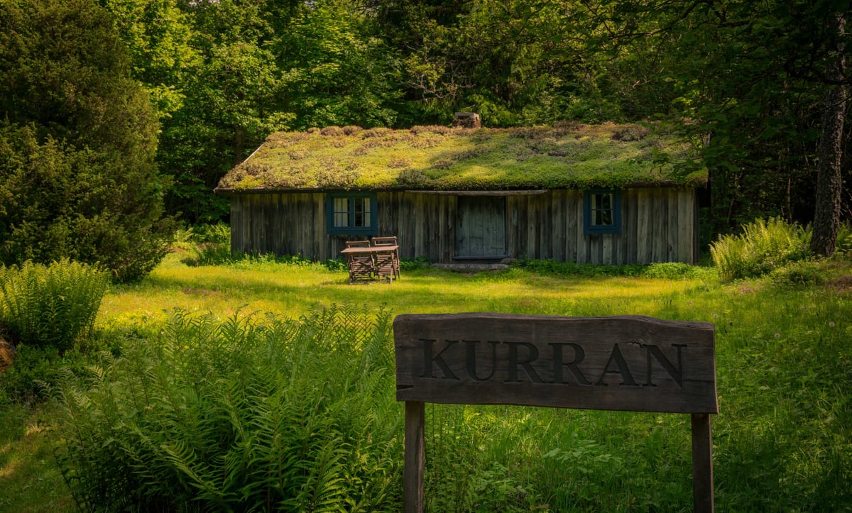 Verweerde houten hut met begroeid grasdak verscholen in een groen bos, met een houten naambordje 'Kurran' op de voorgrond.