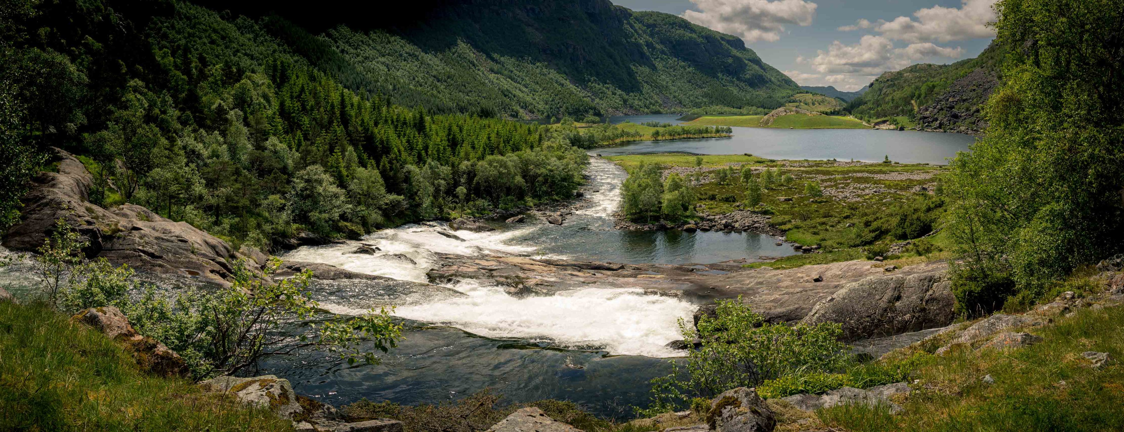 Panoramisch zomers uitzicht op een bruisende rivier met stroomversnellingen in een groene bergvallei met een meer op de achtergrond.