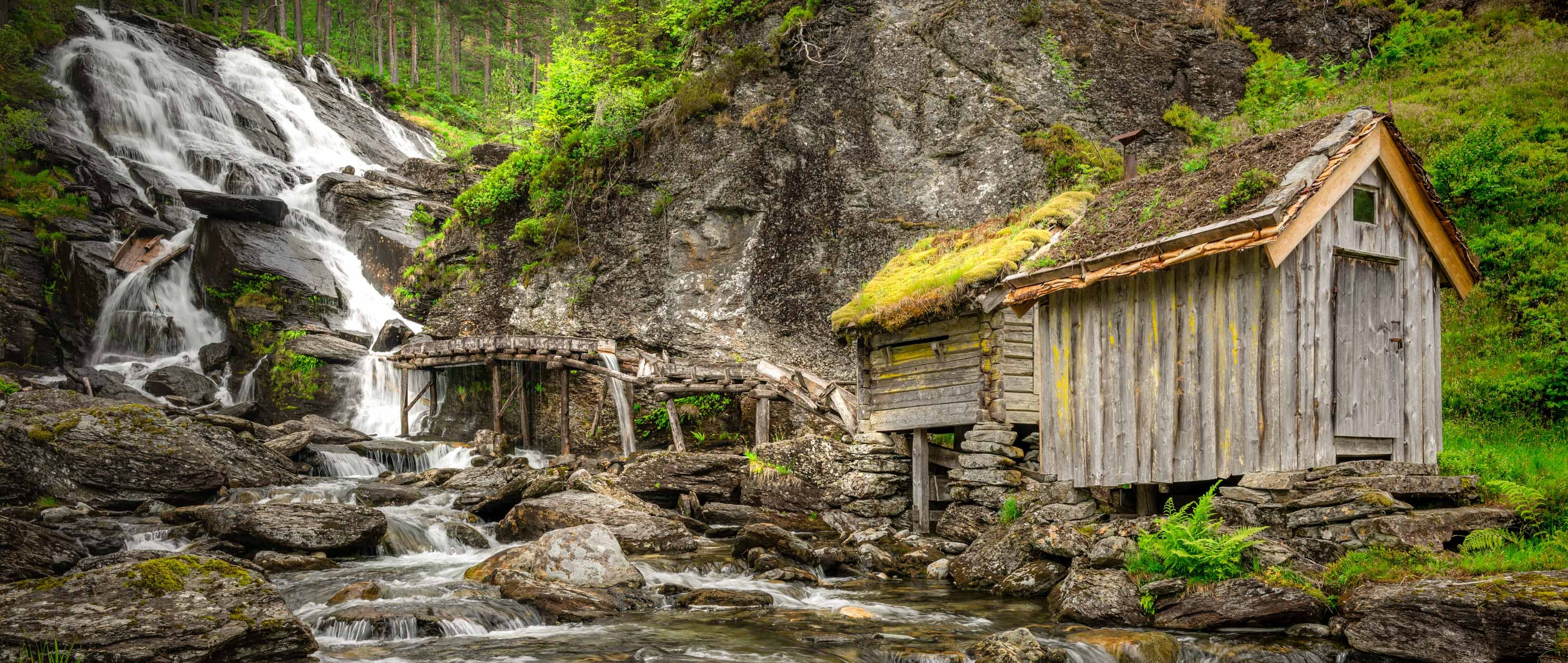 Verlaten houten molen op stenen pijlers naast een waterval in een groen Scandinavisch berglandschap.