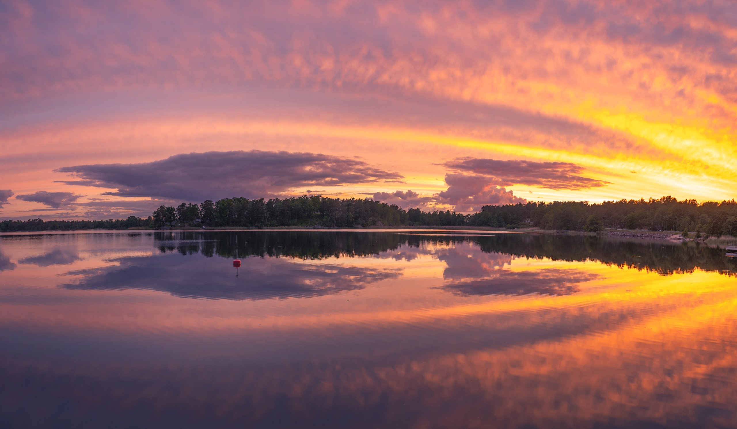 Spiegelgladde baai bij zonsondergang met een vlammende oranje-roze lucht die perfect wordt gereflecteerd in het water, omringd door boomsilhouetten.