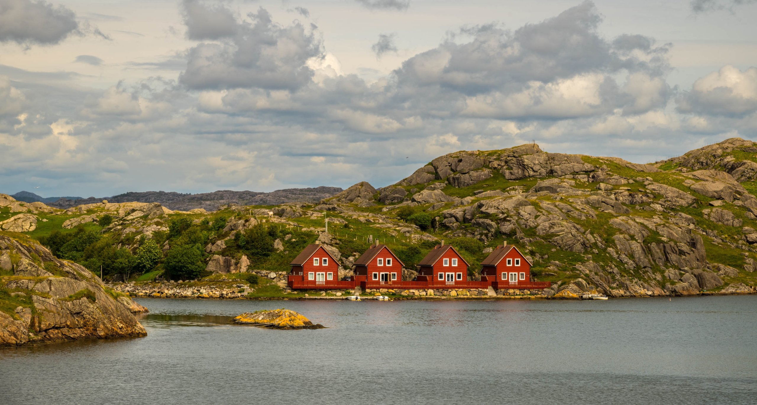Drie rode Noorse houten huizen aan de oever van een fjord, omgeven door rotsachtig landschap onder een bewolkte lucht.