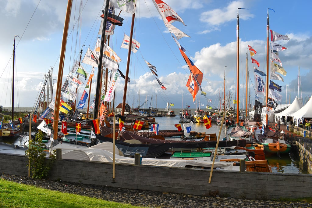 Drukke jachthaven met traditionele houten zeilboten vol wapperende sponsor- en seinvlaggen aan hoge masten onder een bewolkte lucht.