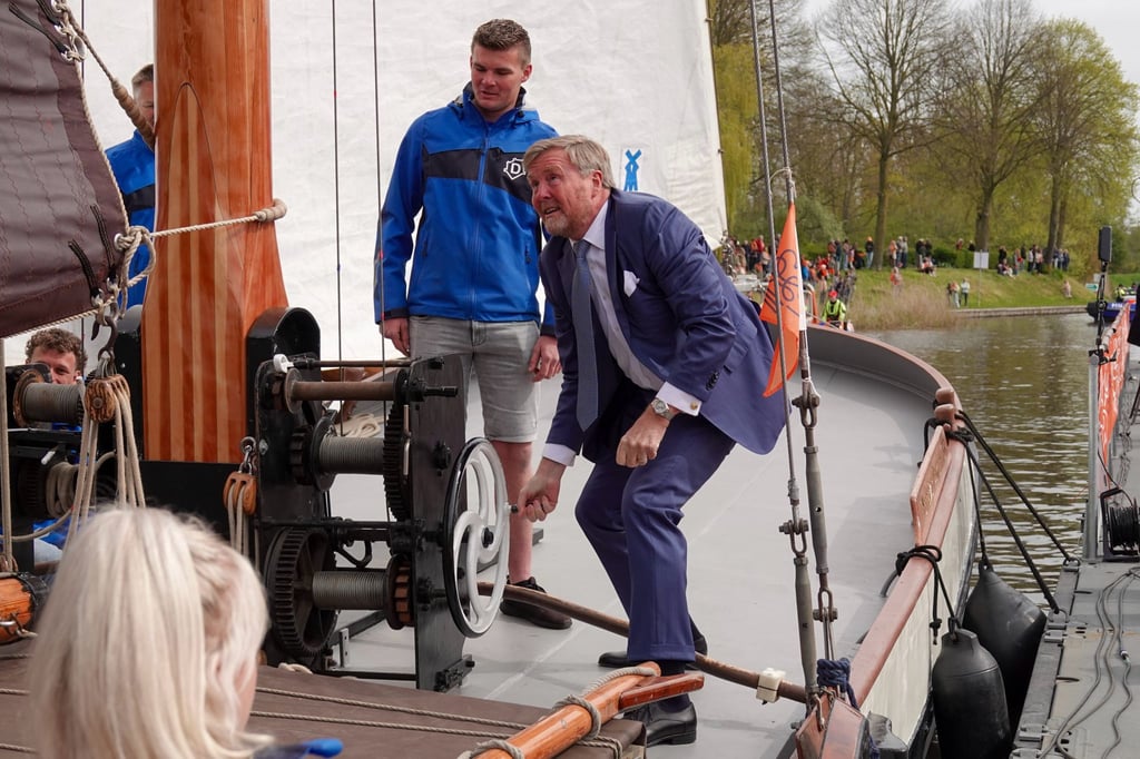 Een man in pak buigt voorover bij het ankermechanisme van een historisch zeilschip terwijl een jongere man in blauw jasje toekijkt.