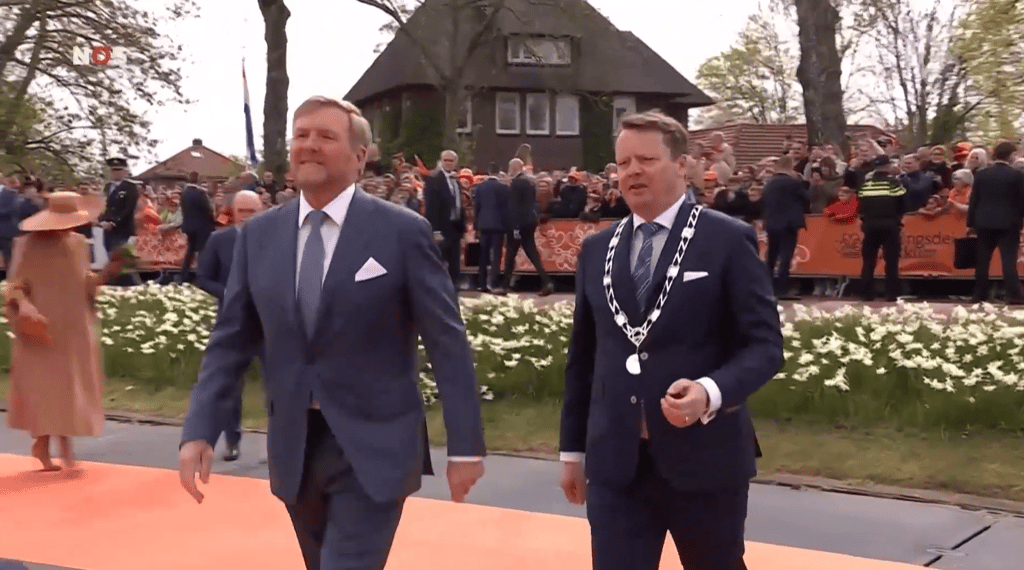Twee mannen in pakken, waaronder een persoon met een burgemeestersketen, lopen over een oranje loper tijdens een officiële Nederlandse feestelijkheid.