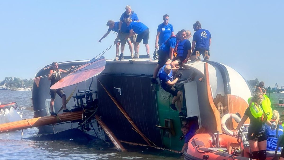 Een gekanteld zeilschip op zijn zij in open water, omringd door reddingsboten en hulpverleners die mensen proberen te redden.