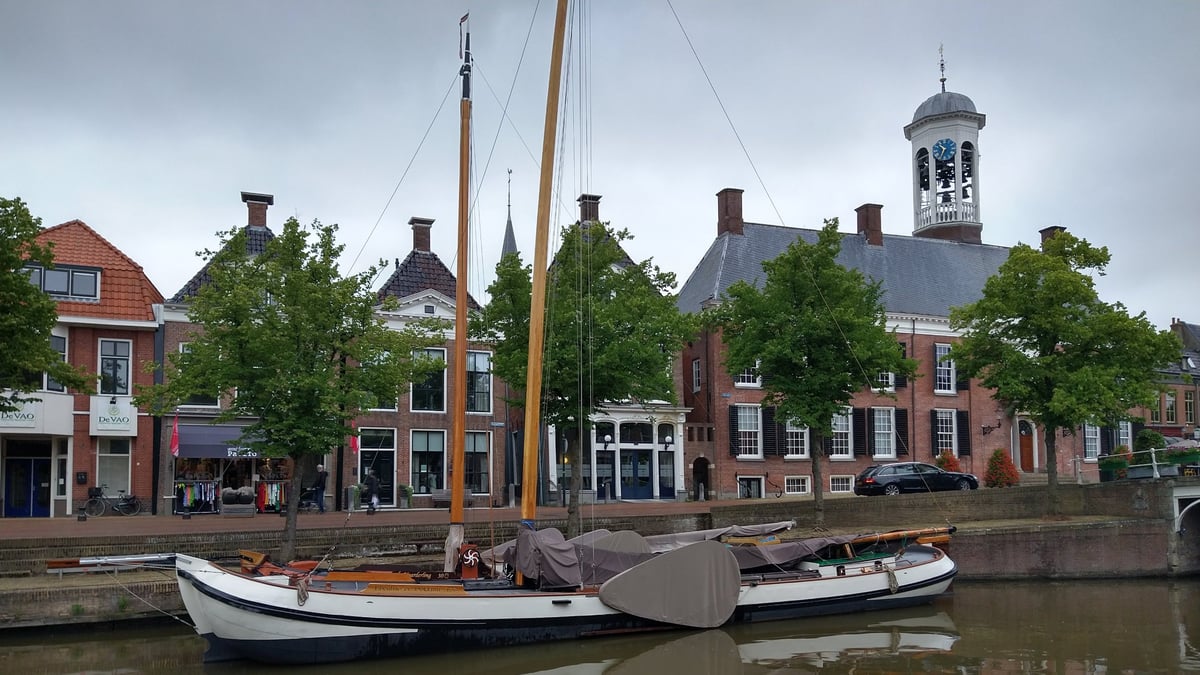 Historisch houten zeilschip afgemeerd in een Nederlandse gracht met 17e-eeuwse bakstenen grachtenpanden en een witte klokkentoren op de achtergrond.
