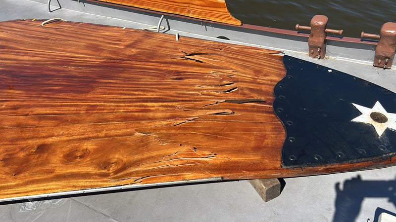 Beschadigde houten romp van een klassieke boot met barsten en een zwart gelakt sterdecor aan de punt, liggend op een steiger.