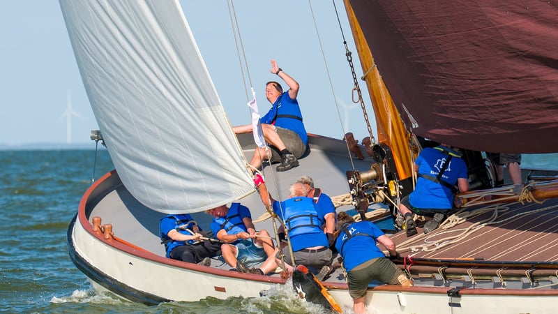 Bemanning in blauwe shirts en zwemvesten werkt samen op de boeg van een klassieke zeilboot tijdens een race op open water.