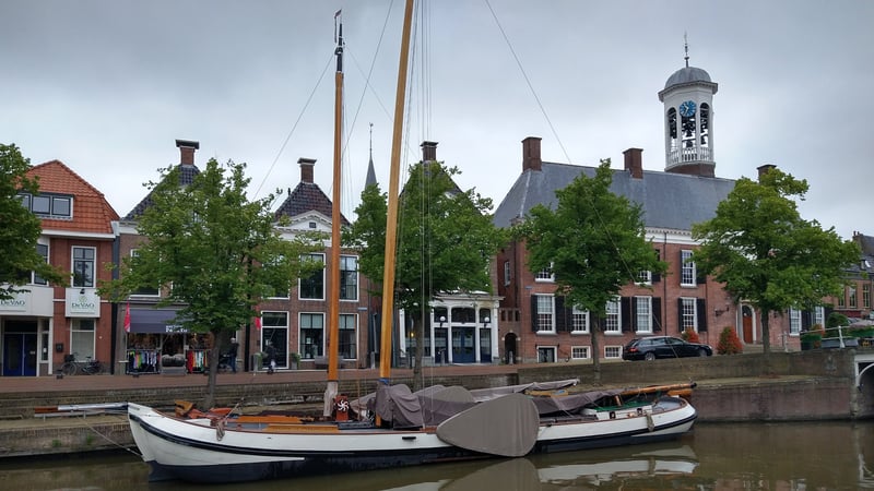 Historisch houten zeilschip afgemeerd in een Nederlandse gracht met 17e-eeuwse bakstenen grachtenpanden en een witte klokkentoren op de achtergrond.