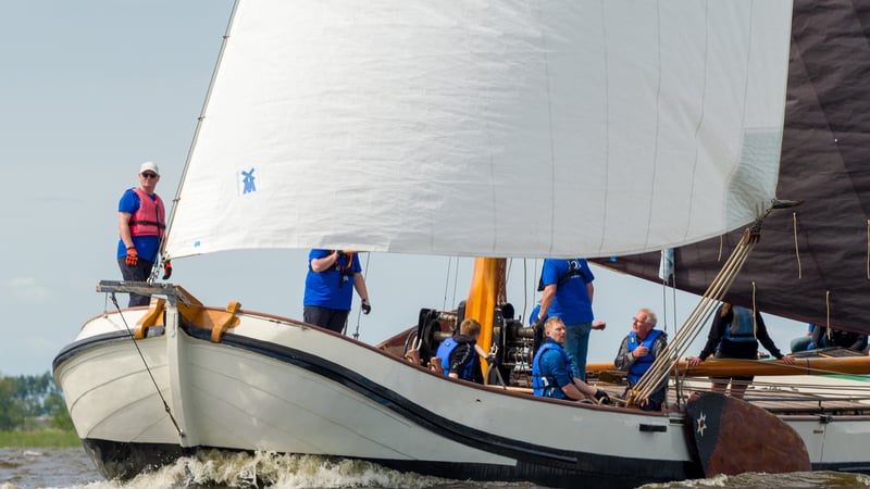 Bemanning in blauwe shirts zeilt op een klassieke witte houten zeilboot over Nederlands binnenwater tijdens een zeilwedstrijd.