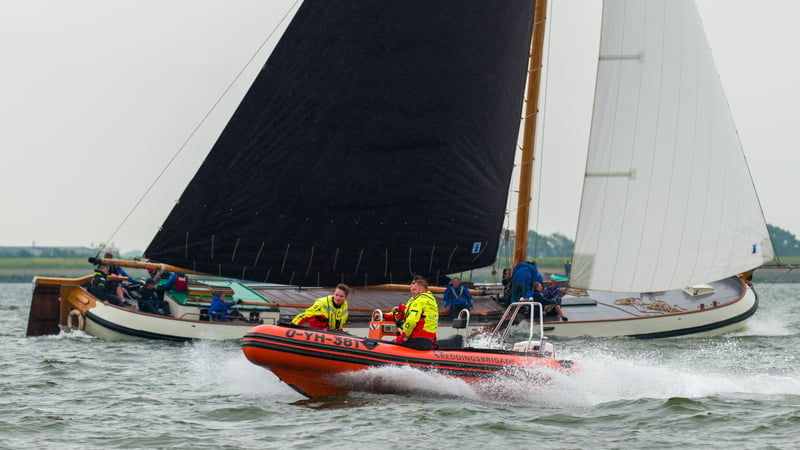 Oranje Reddingsbrigade-rubberboot met twee bemanningsleden in gele jassen vaart snel voor een klassiek houten zeilschip met zwarte zeilen.